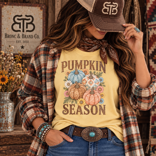 A woman models a yellow graphic t-shirt with a 'Pumpkin Season' design, styled with a brown plaid flannel shirt, a bandana, jeans, and a cap. A 'Bronc & Brand Co.