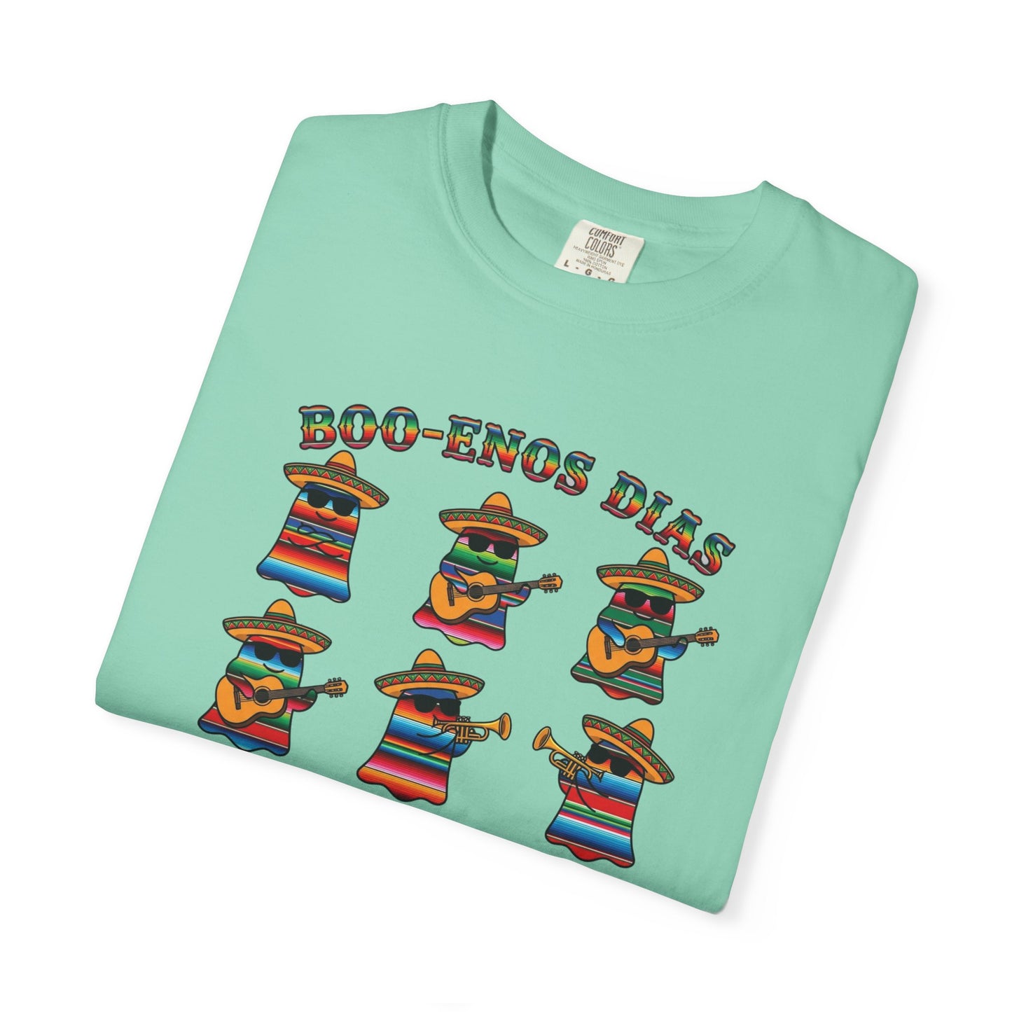 A folded mint green t-shirt on a white background, featuring a colorful graphic of six cartoon characters dressed in traditional Mexican attire playing musical instruments under the text 'BOO-ENOS DIAS'.