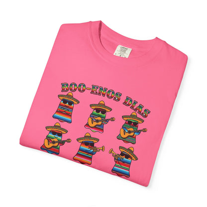 A folded pink t-shirt on a white background, featuring a colorful graphic of six cartoon characters in sombreros playing musical instruments under the text 'BOO-ENOS DIAS'.