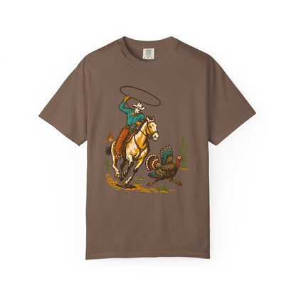 A brown short-sleeve crewneck t-shirt featuring a western-themed graphic print of a cowboy on a galloping horse trying to rope a turkey, displayed on a clean white background.
