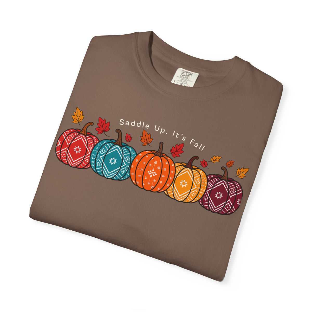 A folded brown crewneck t-shirt featuring a graphic of five patterned pumpkins in red, blue, orange, and purple, with small autumn leaves and the text 'Saddle Up, It's Fall' printed on the chest.