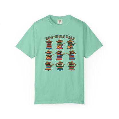 A mint green short-sleeve t-shirt with a colorful graphic print on the front. The graphic features the text 'BOO-TENOS DIAS' above nine cartoon characters wearing sombrero and colorful striped serapes, posed in different dancing stances.