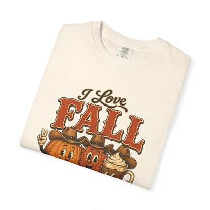 A folded cream-colored t-shirt with a graphic print that reads 'I Love FALL' in brown and orange lettering, featuring cartoon pumpkins wearing cowboy hats and a hot chocolate mug on a white background.