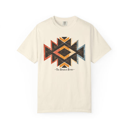 An off-white short-sleeve t-shirt featuring a distressed geometric Southwestern-style diamond pattern in black, orange, and red on the chest, with the text '-The Brushed Bronc-' printed below.