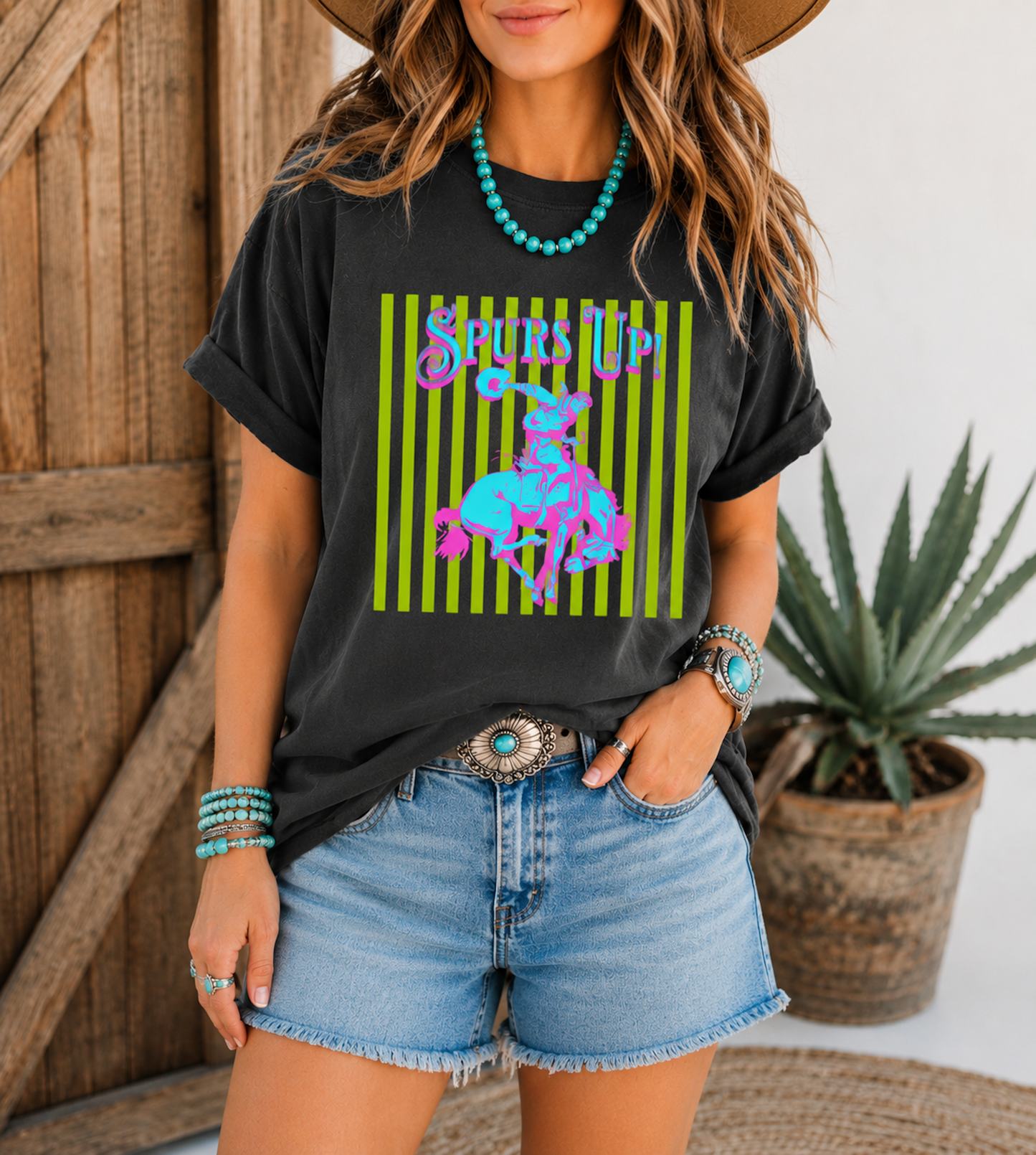 Woman wearing a Periwinkle Comfort Colors Spurs Up graphic tee featuring a colorful retro bronc rider design styled with turquoise jewelry and denim shorts in front of rustic barn doors.