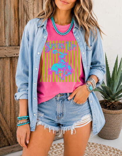 Woman wearing a Periwinkle Comfort Colors Spurs Up graphic tee featuring a colorful retro bronc rider design styled with turquoise jewelry and denim shorts in front of rustic barn doors.