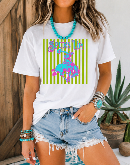 Woman wearing a Periwinkle Comfort Colors Spurs Up graphic tee featuring a colorful retro bronc rider design styled with turquoise jewelry and denim shorts in front of rustic barn doors.