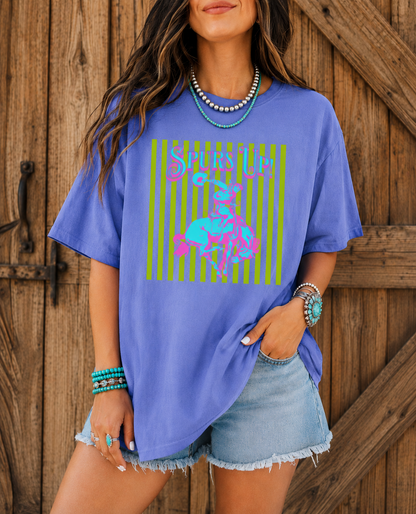Woman wearing a Periwinkle Comfort Colors Spurs Up graphic tee featuring a colorful retro bronc rider design styled with turquoise jewelry and denim shorts in front of rustic barn doors.