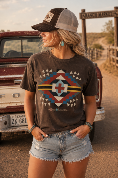 A woman wearing a dark brown graphic t-shirt with a colorful geometric design, paired with denim shorts and a trucker hat, standing in front of a vintage Ford truck on a dirt road.