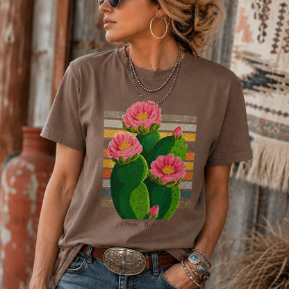 A woman wearing a brown graphic t-shirt with a colorful print of pink cactus flowers, styled with jeans, a western belt, and layered necklaces in a rustic outdoor setting.