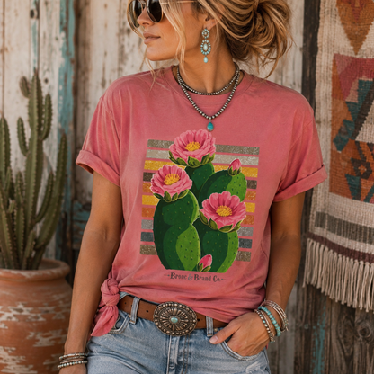A woman wearing a dusty rose graphic t-shirt with a colorful cactus and pink flowers print, paired with blue jeans and western accessories, standing in front of a rustic wooden wall.