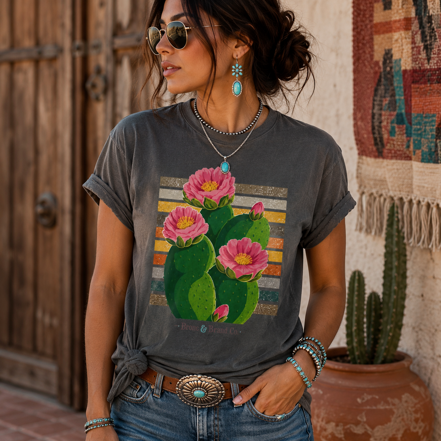 A woman wearing a dark gray graphic t-shirt with a colorful print of pink cactus flowers, styled with blue jeans and accessories in a rustic outdoor setting.