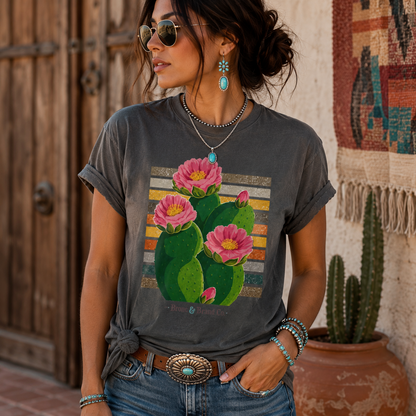 A woman wearing a dark gray graphic t-shirt with a colorful print of pink cactus flowers, styled with blue jeans and accessories in a rustic outdoor setting.