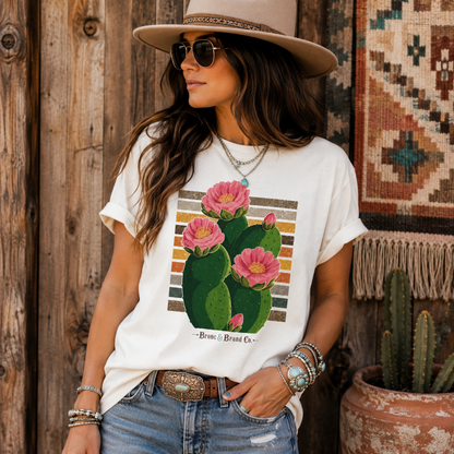 A woman wearing a white graphic t-shirt with a pink cactus print, styled with a wide-brimmed hat, sunglasses, and jeans in front of a rustic wooden fence.