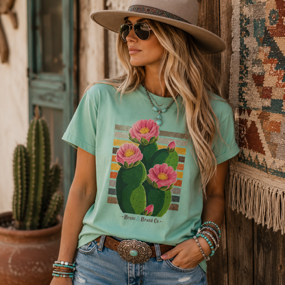 A woman wearing a mint green graphic t-shirt with a pink cactus print, styled with a wide-brimmed hat, sunglasses, and jeans in a rustic outdoor setting.