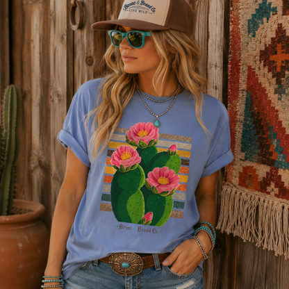 A woman wearing a light blue graphic T-shirt with a pink cactus print, styled with a brown trucker hat, turquoise sunglasses, and jeans, leaning against a rustic wooden wall.