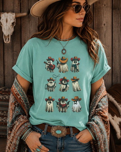 A woman wearing a light aqua graphic t-shirt with a western ghost print, styled with a brown cowboy hat, sunglasses, a patterned cardigan, and jeans against a rustic wooden wall.