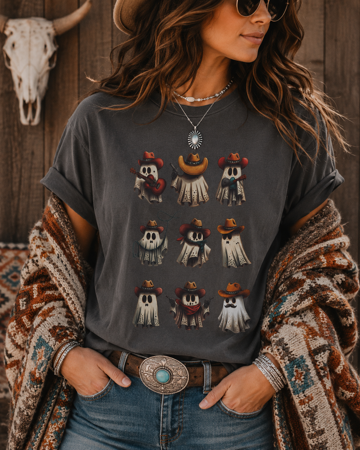 A woman wearing a dark gray graphic t-shirt with a grid of cartoon ghosts in cowboy hats, styled with a bohemian knit shawl, jeans, and a western belt against a rustic wooden wall.