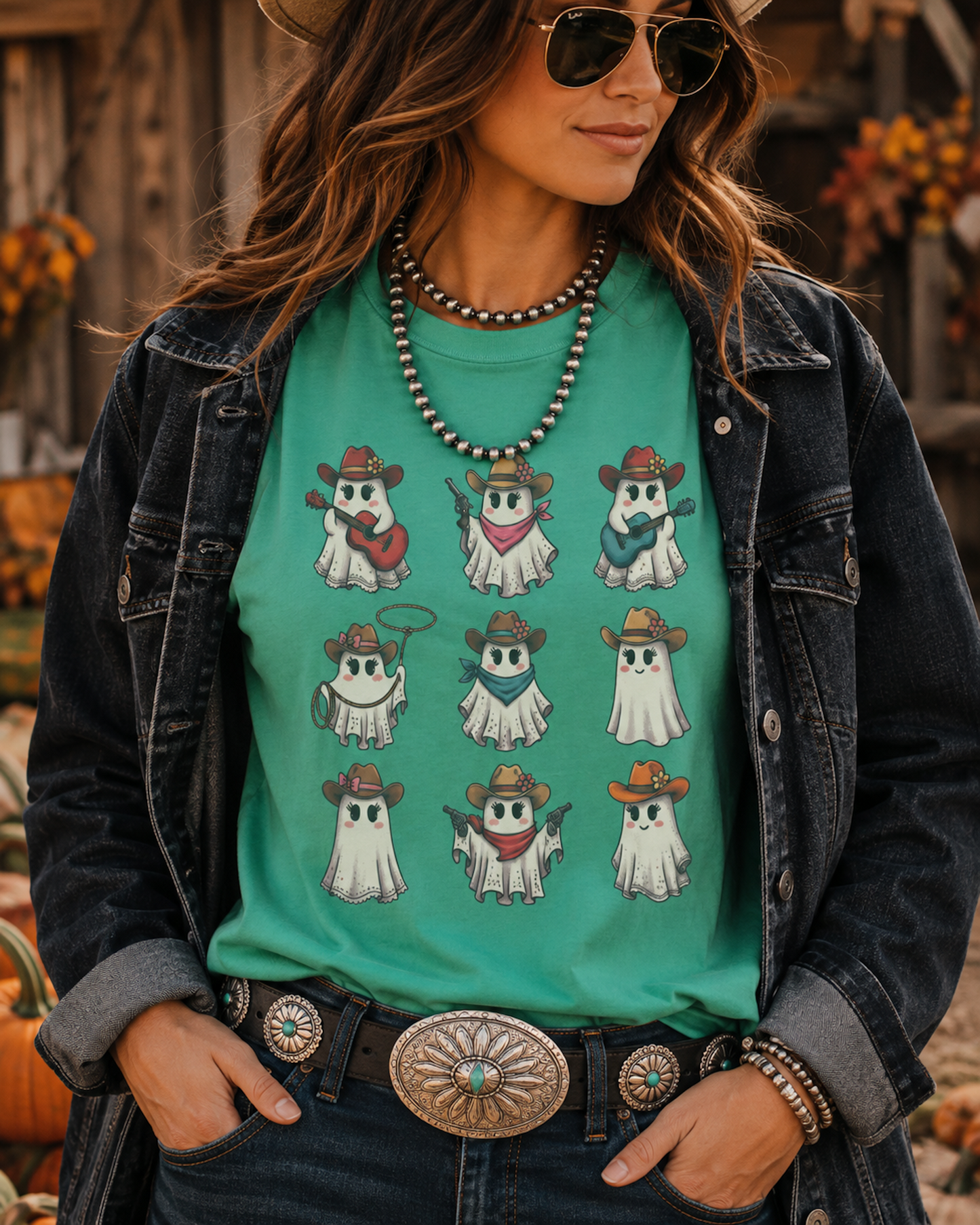 A woman wearing a teal graphic t-shirt with cartoon ghosts dressed as cowboys, styled with a dark denim jacket, jeans, and a western belt in an autumn-themed setting.
