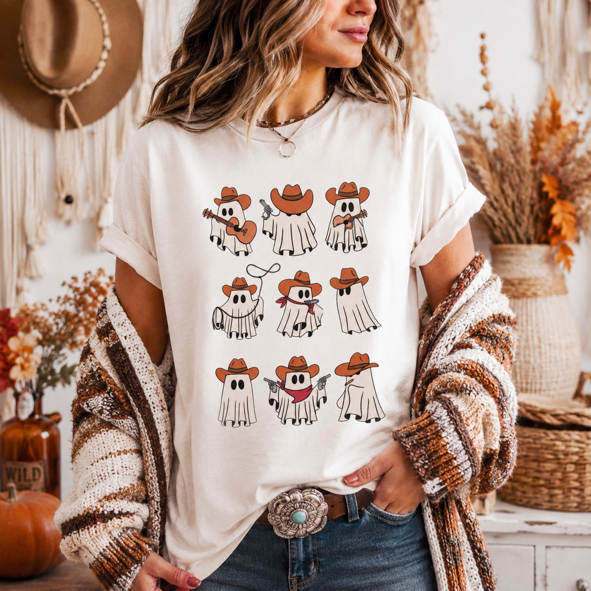 A woman wearing a cream-colored graphic t-shirt with a cartoon ghost cowboy print, styled with a chunky knit brown and white cardigan, blue jeans, and a large western-style belt buckle. The background is a cozy, warm-toned rustic room with a macrame wall hanging and dried flowers.