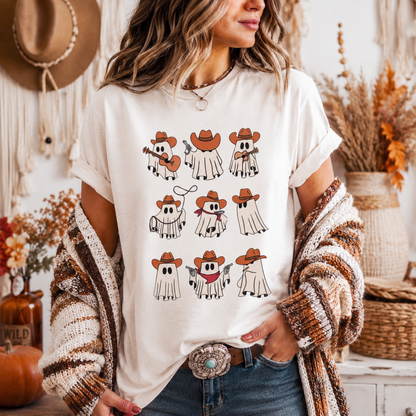 A woman wearing a cream-colored graphic t-shirt with a cartoon ghost cowboy print, styled with a chunky knit brown and white cardigan, blue jeans, and a large western-style belt buckle. The background is a cozy, warm-toned rustic room with a macrame wall hanging and dried flowers.