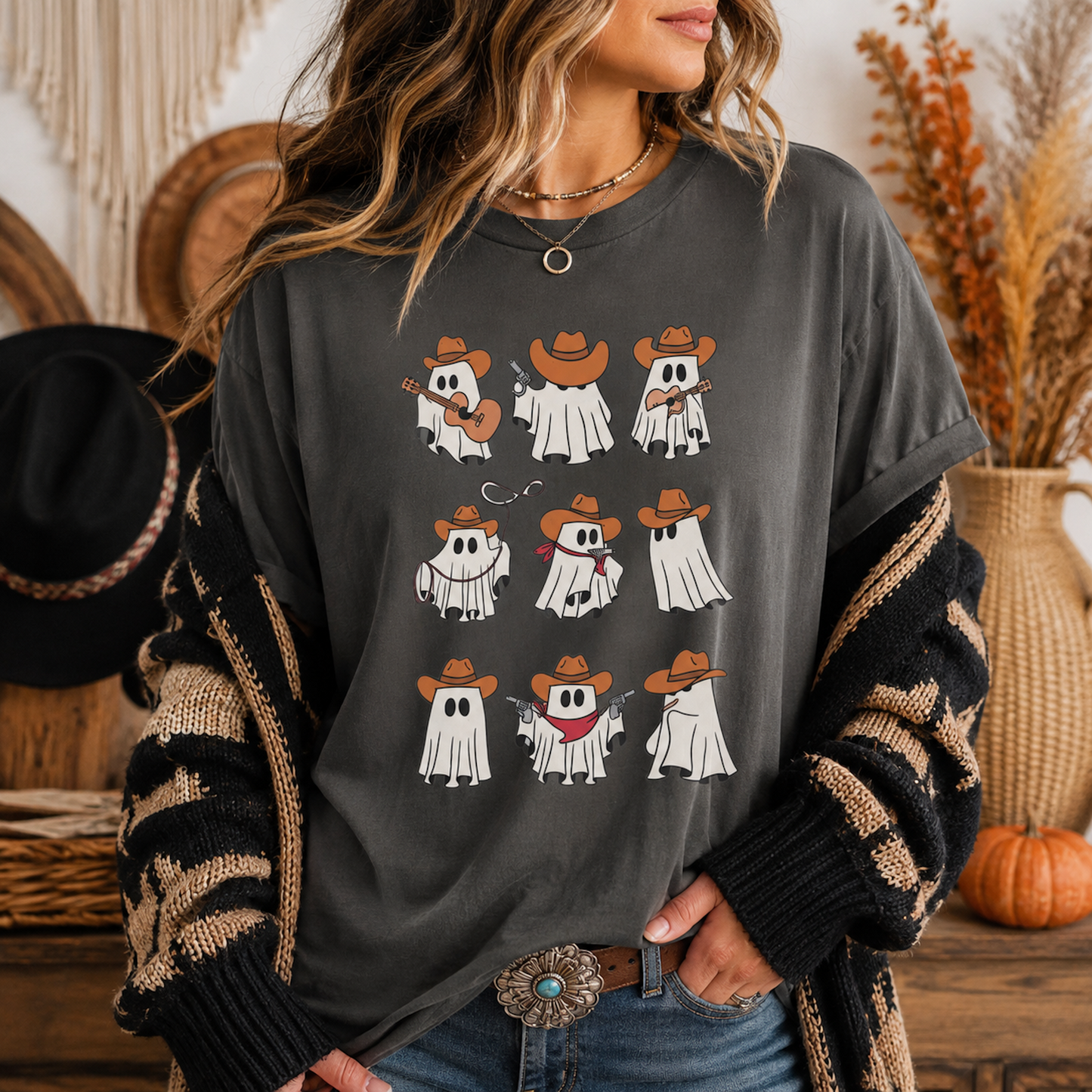 A woman wearing a dark gray graphic t-shirt with a 3x3 grid of cute cartoon ghosts in cowboy hats, styled with a black and beige knit cardigan, blue jeans, and a western belt buckle in a cozy autumn-themed room.