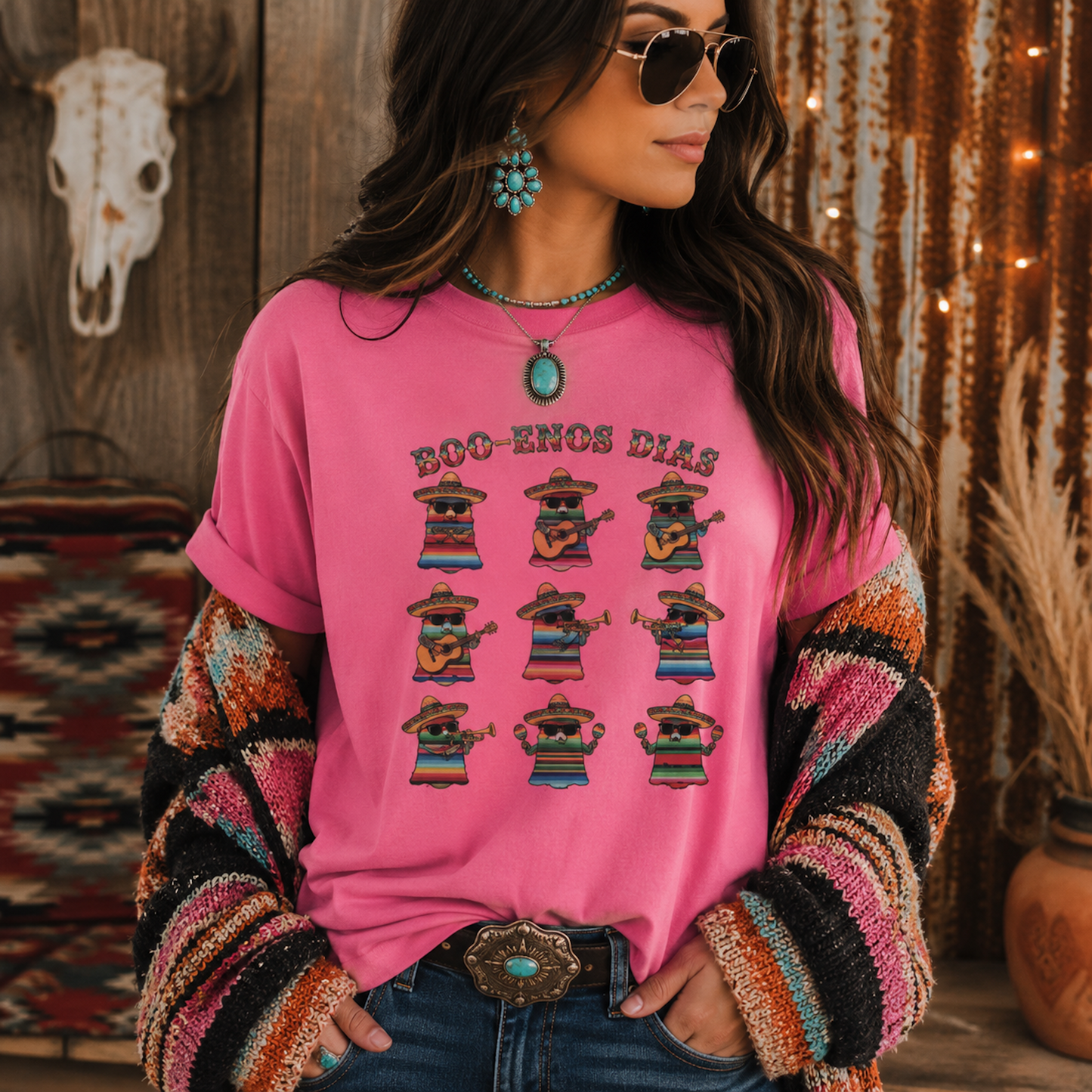 A woman wearing a bright pink graphic t-shirt with 'BOO-ENOS DIAS' and ten illustrated figures in colorful ponchos, styled with a striped knit cardigan, jeans, turquoise jewelry, and sunglasses in a rustic bohemian setting.