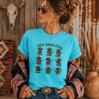 A woman wearing a bright turquoise graphic T-shirt with the text 'BOO-ENOS DIAS' and a row of colorful mariachi-style hat graphics, styled with a striped cardigan, jeans, and sunglasses in a rustic indoor setting.