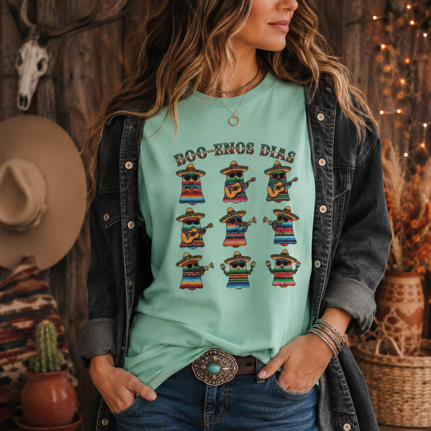 A woman wearing a mint green graphic t-shirt with 'BOO-ENOS DIAS' and colorful dancing figures, styled with a dark denim jacket and jeans in a rustic, warmly lit room.