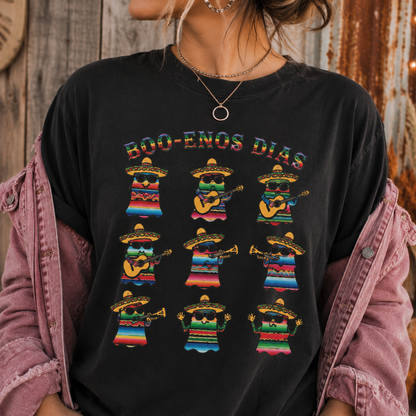 A person wearing a black graphic T-shirt with the text 'BOO-ENOS DIAS' and nine colorful illustrated figures in striped clothing, styled with a pink denim jacket draped off the shoulders against a rustic wooden background.