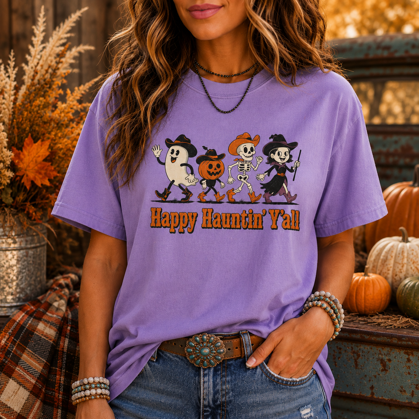A woman wearing a lavender purple graphic t-shirt with a 'Happy Haunting' Halloween print, styled with blue jeans and accessories in a rustic autumn setting.