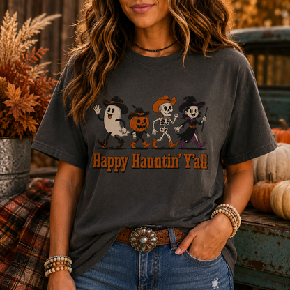A woman wearing a dark charcoal gray graphic t-shirt with a 'Happy Haunting' Y'all Halloween print, paired with blue jeans and a brown belt, standing in a rustic autumn-themed setting.