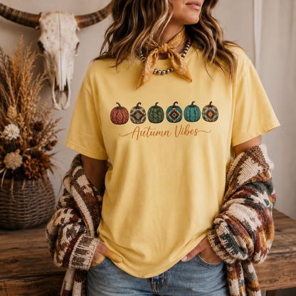 A woman wearing a pale yellow graphic T-shirt with five patterned pumpkins and the words 'Autumn Vibes' across the chest, styled with a tan bandana, beaded necklace, and a multicolor knit cardigan in a rustic fall-themed room.