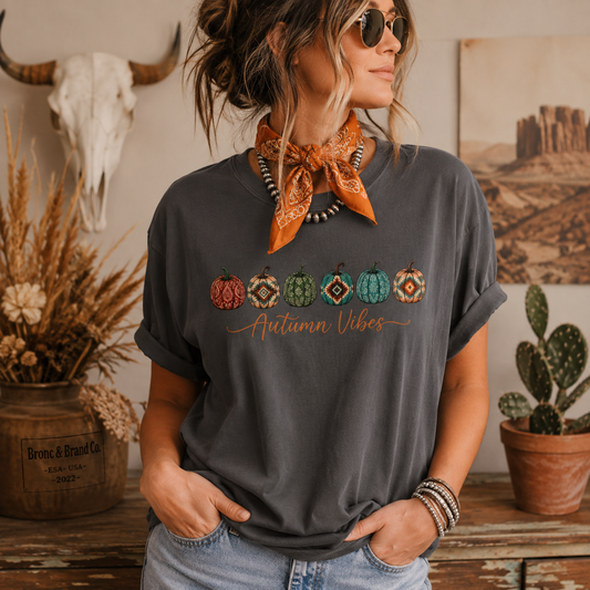 A woman wearing a dark charcoal gray graphic t-shirt with a colorful autumn pumpkin design and 'Autumn Vibes' text. She is styled with an orange paisley neck bandana, beaded necklaces, and sunglasses, standing in a rustic, western-themed room with a wooden table, potted cactus, and a bull skull on the wall.