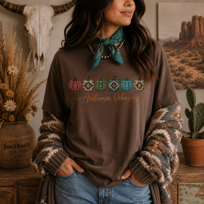 A woman wearing a brown graphic t-shirt with a colorful pumpkin design and 'Autumn Vibes' text, styled with a patterned bandana, knit cardigan, and jeans in a rustic indoor setting.