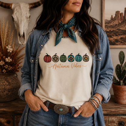 A woman wearing a cream-colored graphic t-shirt with a western-themed autumn design, styled with a denim shirt, a teal bandana, and jeans in a rustic room setting.