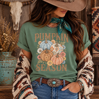 A woman wearing a sage green graphic t-shirt with a 'Pumpkin Season' design, styled with a brown cowboy hat, a teal bandana, a chunky knit cardigan, and blue jeans. The brand 'Bronte & Brand Co' is visible on a rustic blue vase in the background.