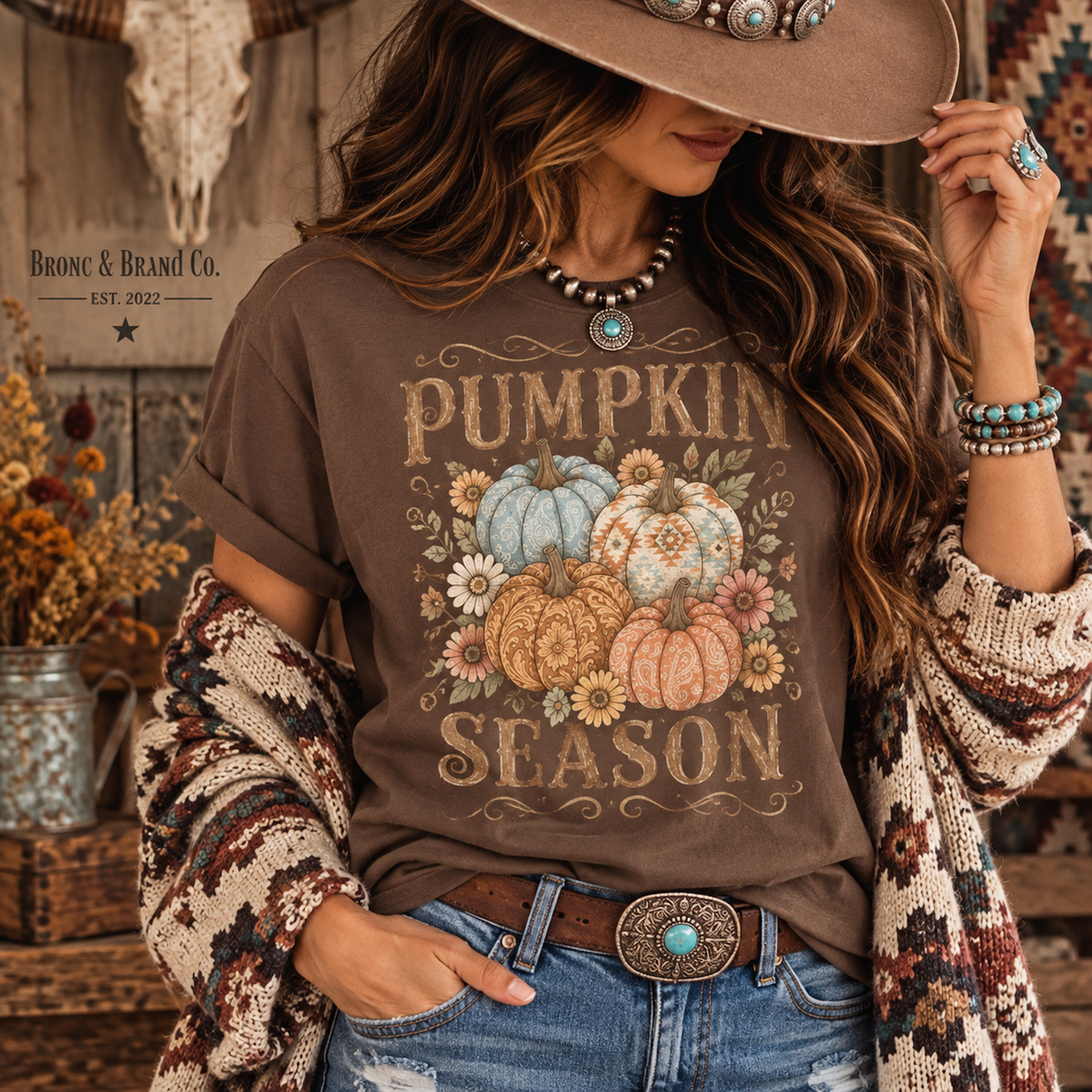 A woman wearing a brown graphic T-shirt with a fall pumpkin design, styled with a wide-brim hat, patterned cardigan, jeans, and turquoise jewelry. The brand name 'Bronc & Brand Co.
