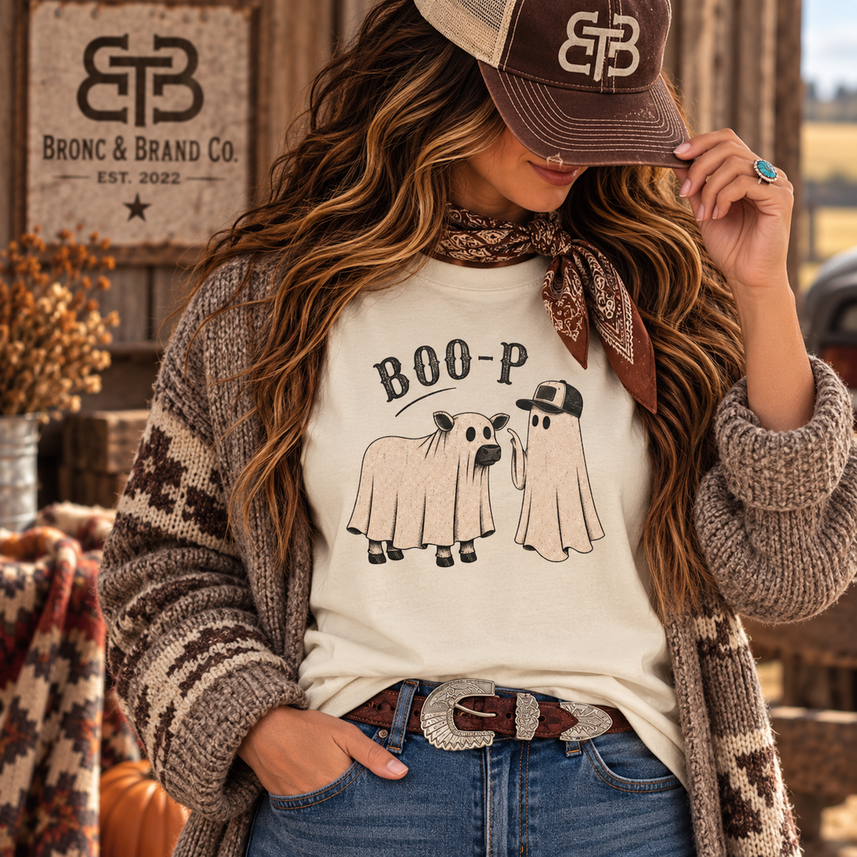 A woman wearing a cream graphic T-shirt with a ghost-themed 'BOO-P' print, styled with a brown patterned cardigan, jeans, and a brown cap. The background shows a rustic wooden sign for 'Bron & Brand Co'.