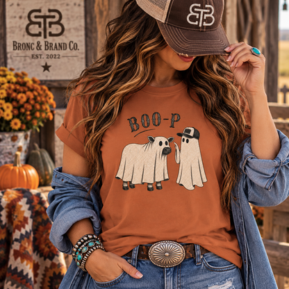 A woman wearing a rust-orange graphic t-shirt with a 'BOO-P' cartoon sheep and ghost design, styled with a denim jacket, jeans, and a brown trucker hat. The background features a rustic wooden sign for 'Bronc & Brand Co.