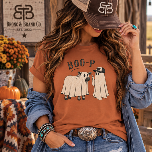 A woman wearing a rust-orange graphic t-shirt with a 'BOO-P' cartoon sheep and ghost design, styled with a denim jacket, jeans, and a brown trucker hat. The background features a rustic wooden sign for 'Bronc & Brand Co.
