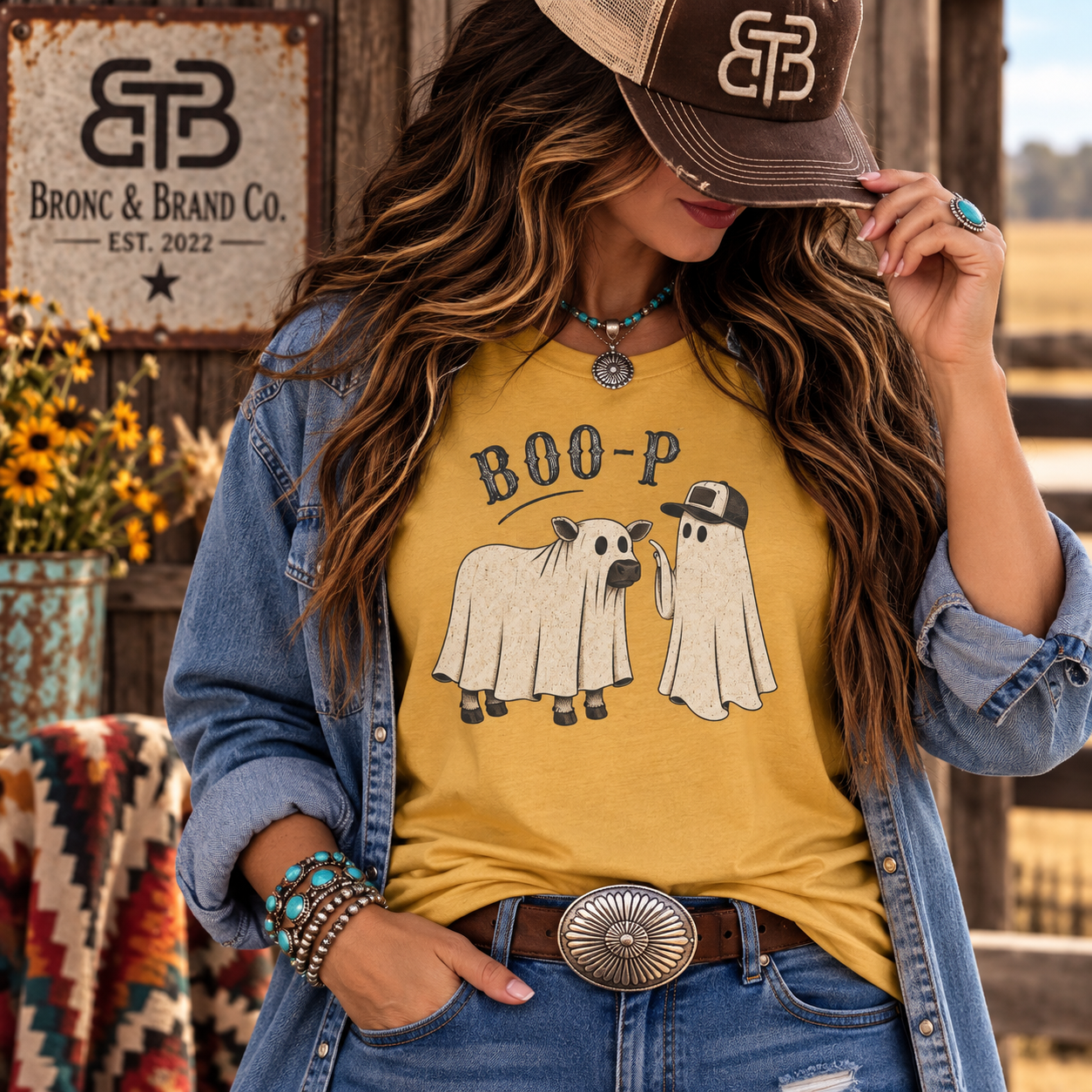 A woman wearing a mustard yellow graphic t-shirt with a 'BOO-P' ghost design, styled with a denim jacket, jeans, and a brown cap in a rustic outdoor setting.