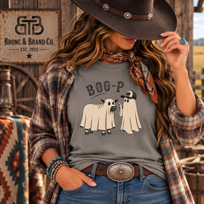 A woman wearing a gray graphic t-shirt with a 'BOO-P' ghost design, styled with a brown cowboy hat, a brown plaid flannel shirt, and blue jeans in a rustic western-themed setting. The brand logo 'Bronc & Brand Co.