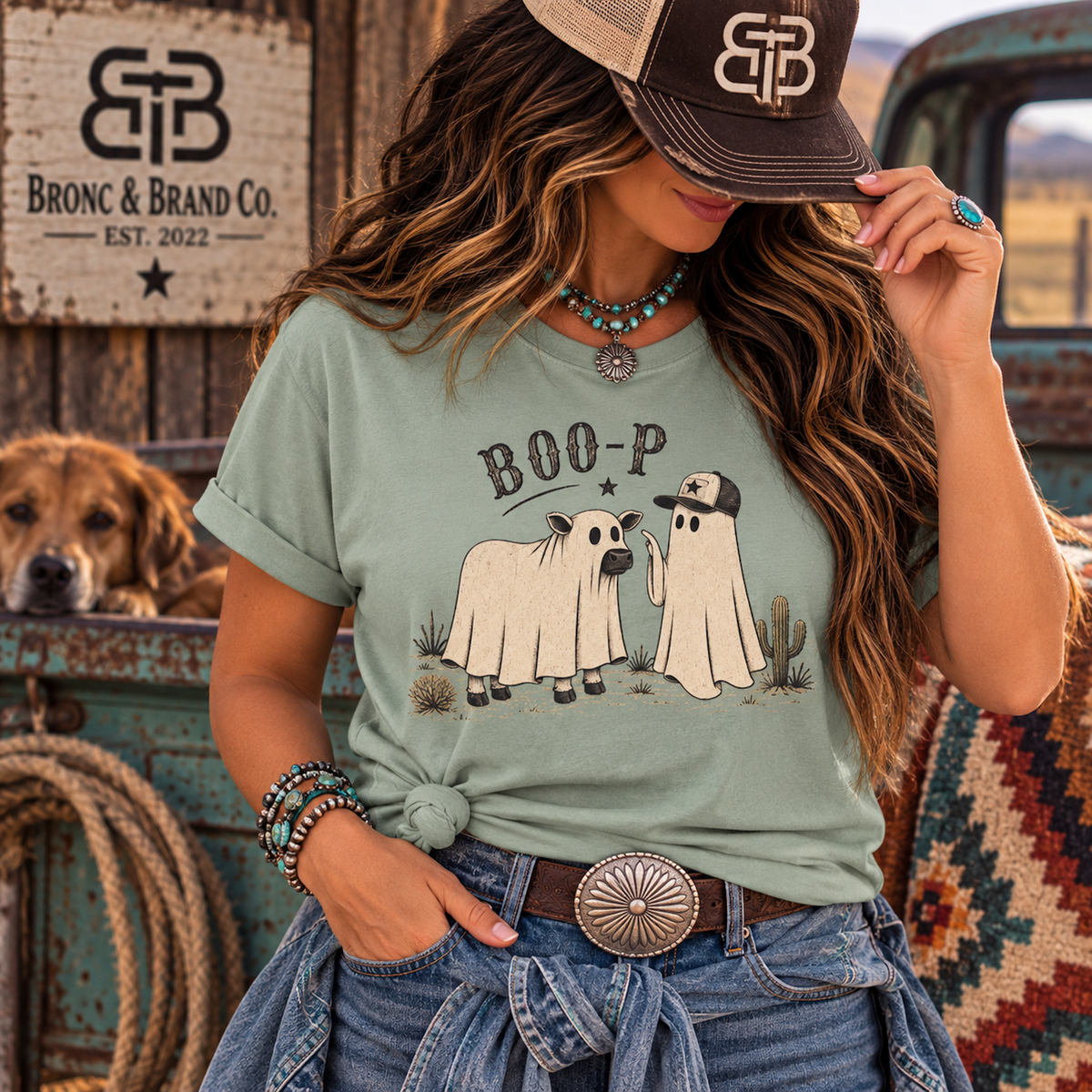 A woman wearing a sage green graphic t-shirt with a 'BOO-P' cartoon ghost dog design, styled with a brown cap, denim jeans, and bracelets. A dog is sitting in the background of a rustic outdoor setting with a wooden sign for 'Bronc & Brand Co.