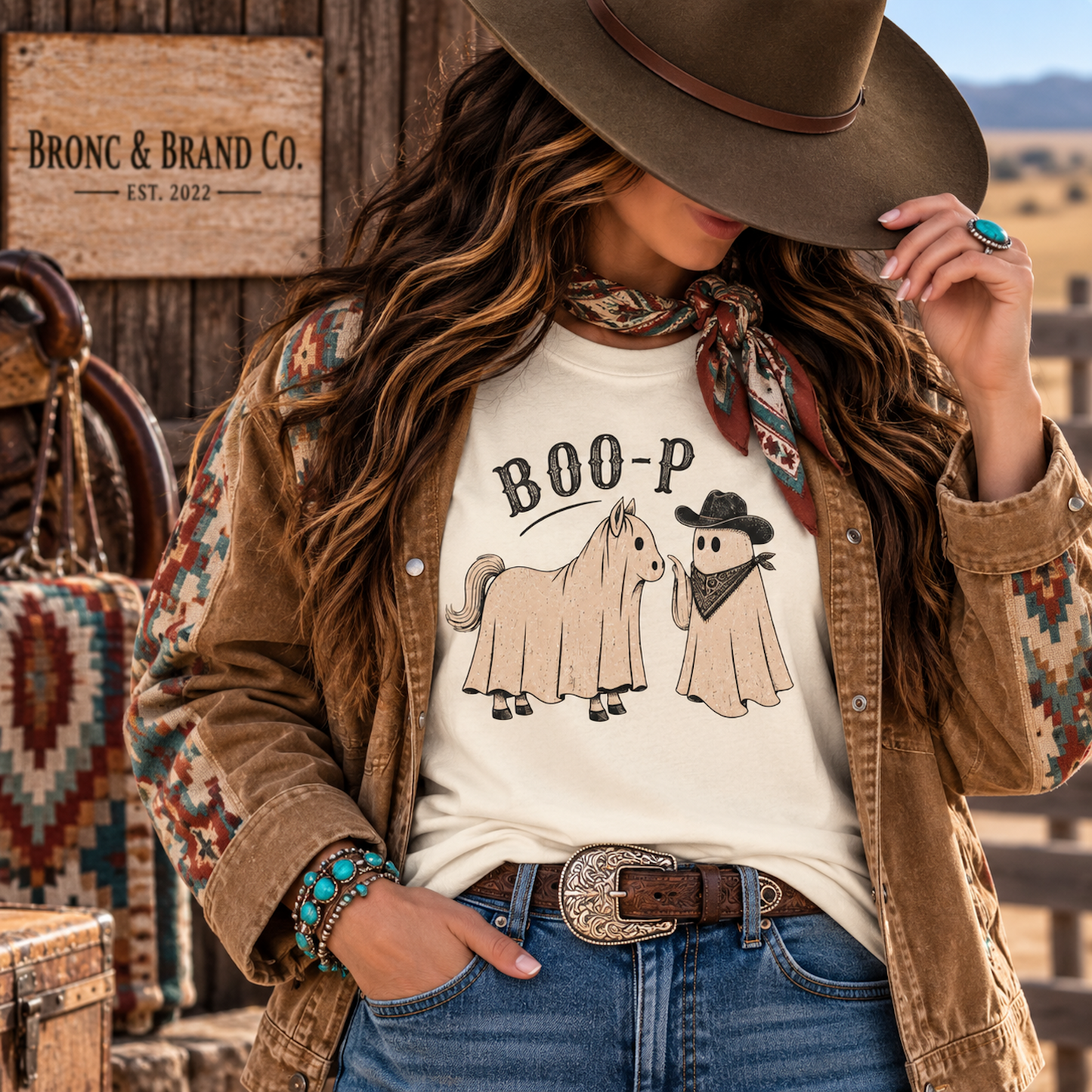 A woman in a rustic western outfit wearing a cream graphic t-shirt with a 'BOO-P' horse and cowboy print, styled with a brown suede cowboy hat, patterned jacket, and jeans. A wooden sign for 'BRONC & BRAND CO.