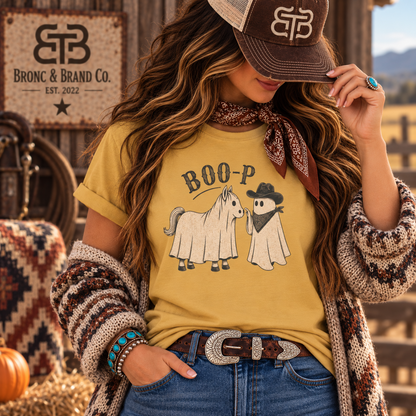 A woman wearing a mustard yellow graphic T-shirt with a ghost horse and ghost cowboy design and the text 'BOO-P', styled with a brown patterned knit cardigan, jeans, and a brown cap. The background shows a rustic barn interior with a sign reading 'EB Bronc & Brand Co.