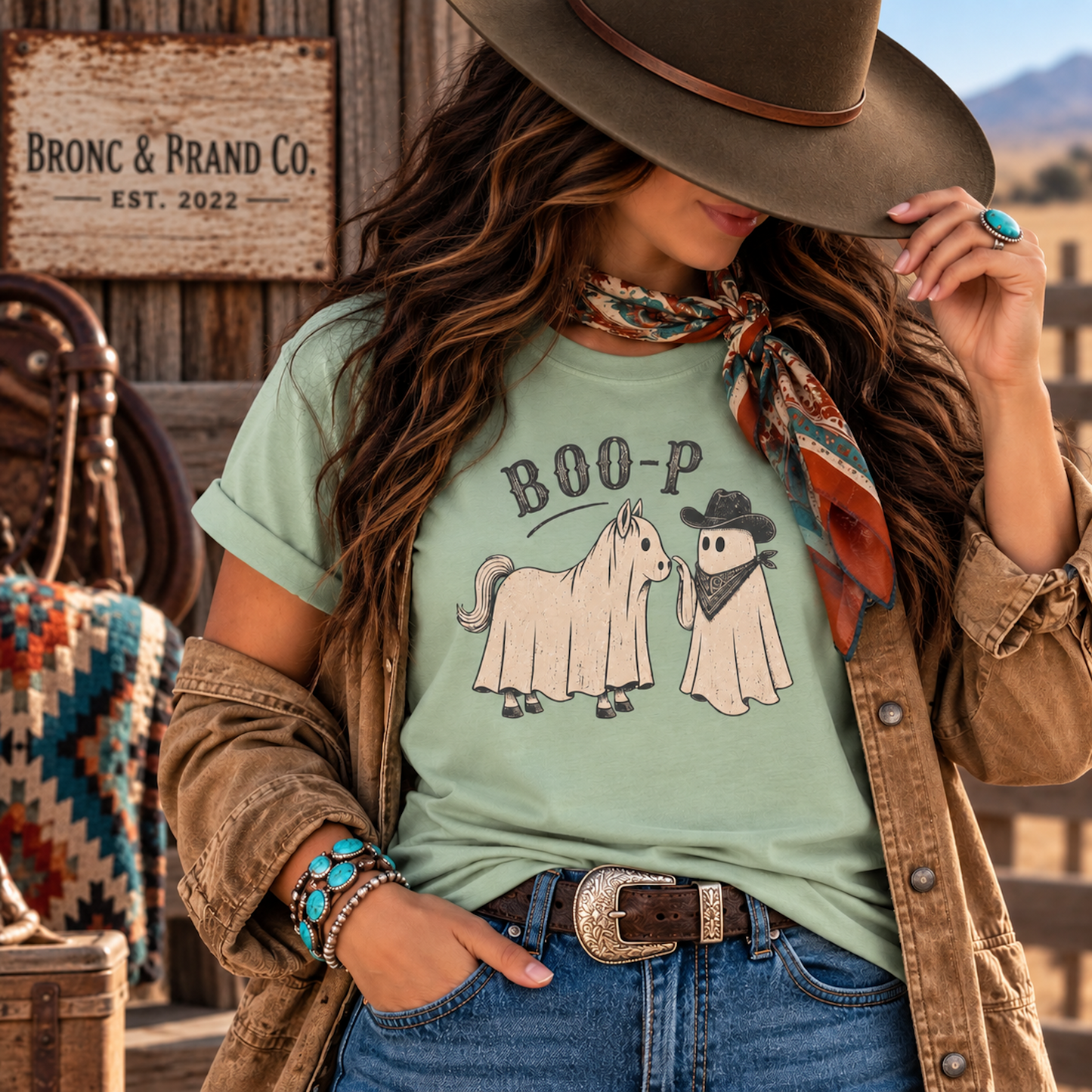 A woman wearing a light green graphic t-shirt with a cowboy and ghost design, styled with a brown jacket, jeans, and a wide-brimmed hat in a rustic western setting.