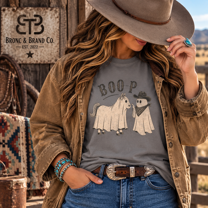 A woman wearing a gray graphic t-shirt with a spooky horse and ghost design, styled with a brown jacket, jeans, and a cowboy hat in a rustic western setting. The brand logo 'Bronc & Brand Co.