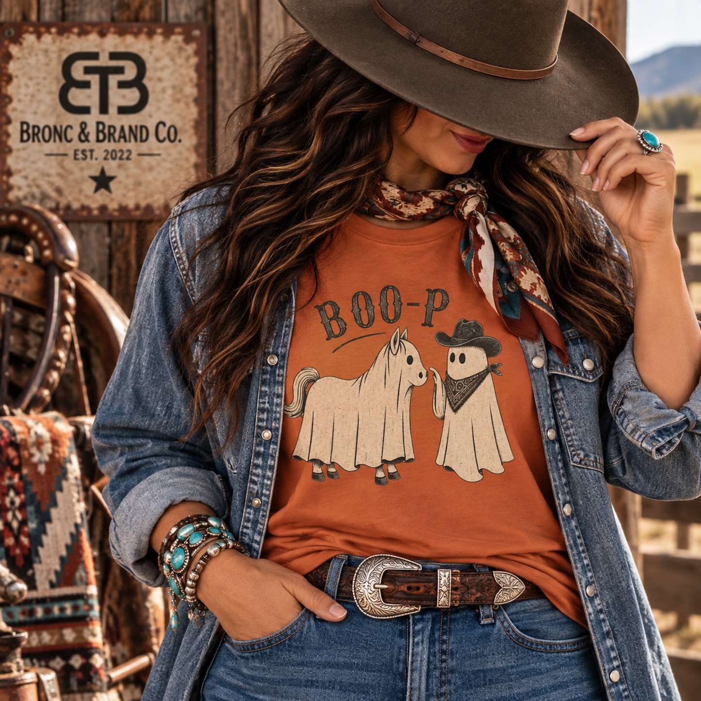 A woman wearing a rust-orange graphic t-shirt with a ghost and horse design and the text 'BOO-P', styled with a denim jacket, a brown wide-brim hat, and a patterned scarf in a rustic western setting. The 'Bronc & Brand Co.