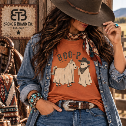 A woman wearing a rust-orange graphic t-shirt with a ghost and horse design and the text 'BOO-P', styled with a denim jacket, a brown wide-brim hat, and a patterned scarf in a rustic western setting. The 'Bronc & Brand Co.